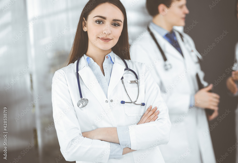 Smiling intelligent woman-doctor is standing with arms crossed in a clinic, together with her colleagues at the background. Portrait of physicians at work. Perfect medical service in a hospital Smiling intelligent woman-doctor is standing with arms crossed in a clinic, together with her colleagues at the background. Portrait of physicians at work. Perfect medical service in a hospital