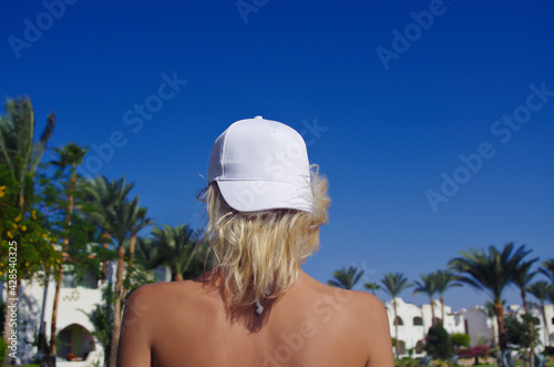 white baseball cap mockup, young beautiful girl with blonde hair in a white baseball cap on a background of blue sky and palm trees