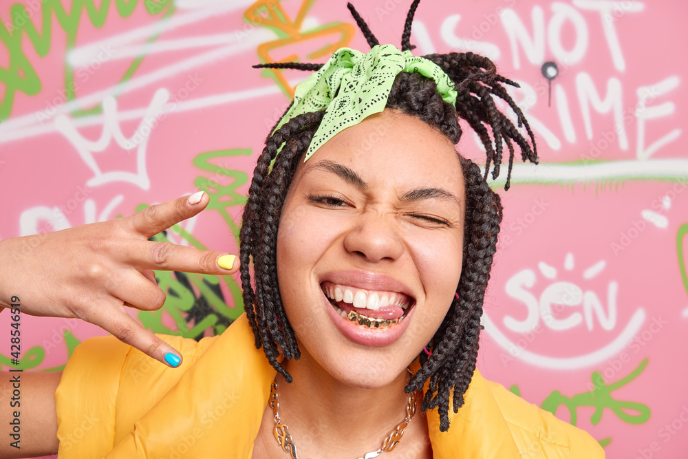 Trendy cheerful hipster girl makes yo gesture shows golden teeth and tongue winks eye enjoys cool modern music stands near graffiti wall enjoys her freedom. Stylish female rapper in urban place - obrazy, fototapety, plakaty 