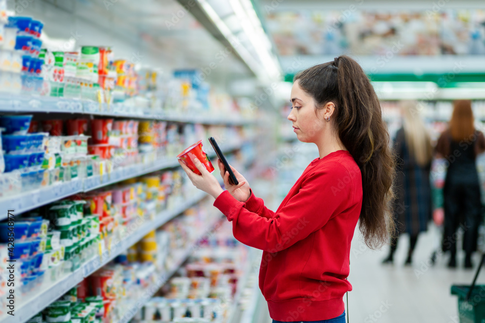 Portrait of young woman scans the QR code on a package of yogurt. In the background, a supermarket with visitors in a blur. Side view. The concept of modern technologies