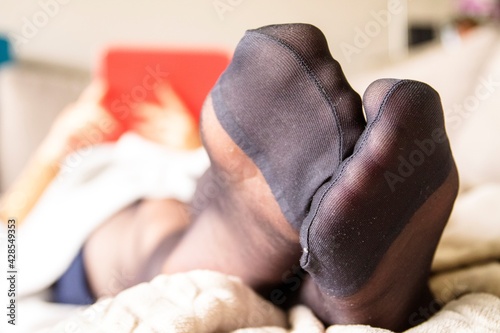 A close up of the black nylon feet of a woman dressed in stockings or pantyhose with reinforced toes, lying on a couch in a living room relaxing and using her tablet computer in the background.