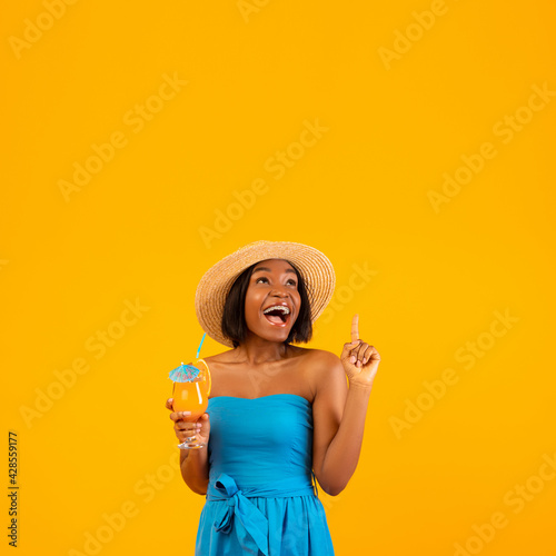 Excited black woman with tasty summer cocktail pointing up at empty space above her head, orange studio background