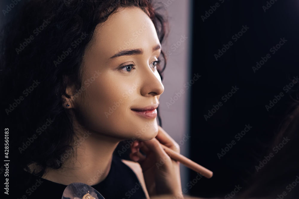 Obraz premium Make up talanted artist doing maquillage to woman applying concealer. Visagist using brushes shadows facial professional cosmetics for makeup. Beauty industry studio, image with copy space.