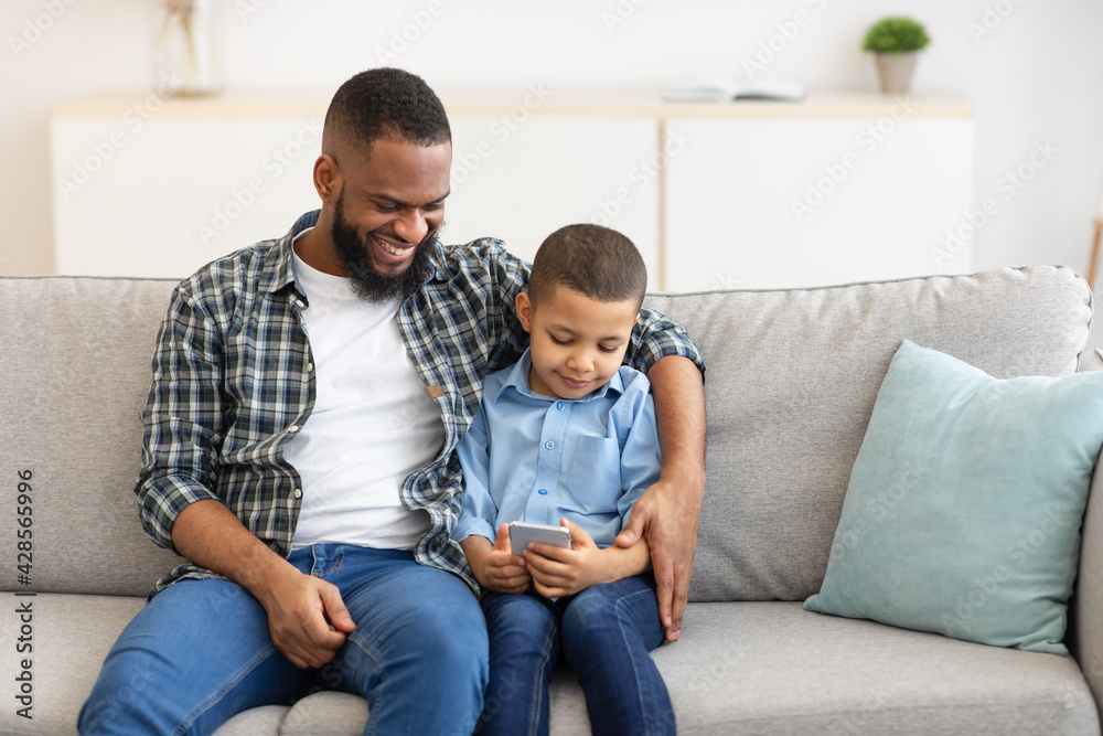 Black Father Hugging Son While He Using Smartphone At Home