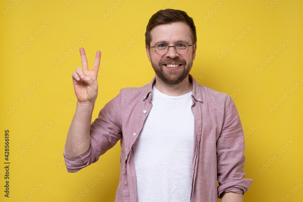 Joyful man in glasses and casual clothes shows the symbol of victory and looks at the camera smiling. Digit with two fingers. Joyful man in glasses and casual clothes shows the symbol of victory and looks at the camera smiling. Digit with two fingers.