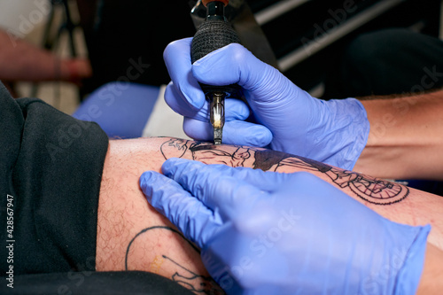 the hands in blue gloves of a tattoo artist doing a tattoo on the leg of a male in a studio