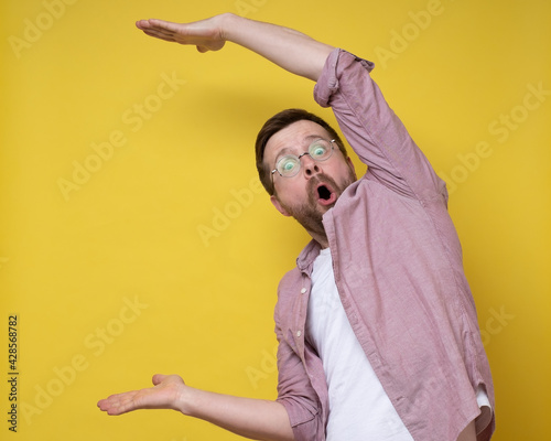Surprised man in glasses shows a large size with hands, he exaggerates and looks at the camera in amazement. Copy space.  photo