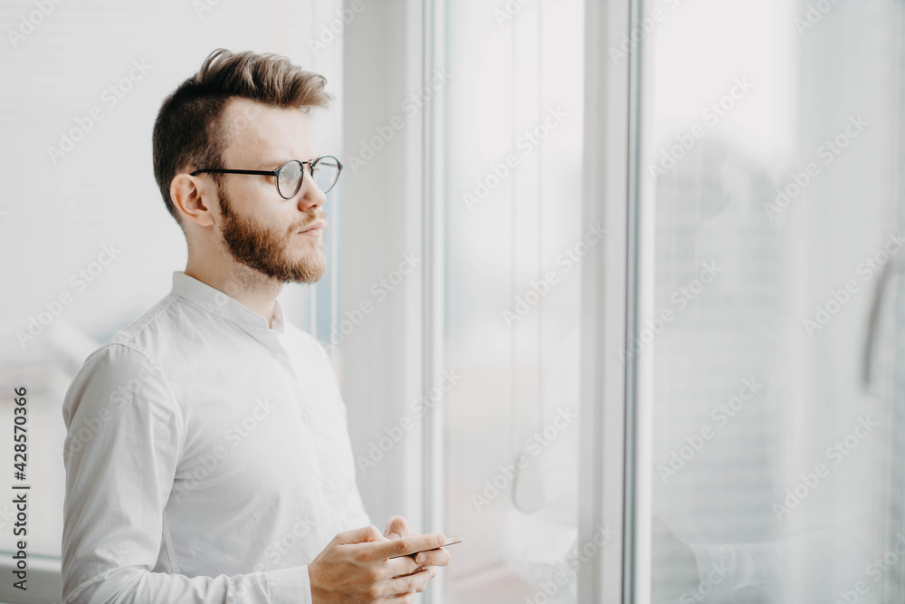 Fototapeta premium man businessman stands by the window and thinks in a white shirt, bearded young man