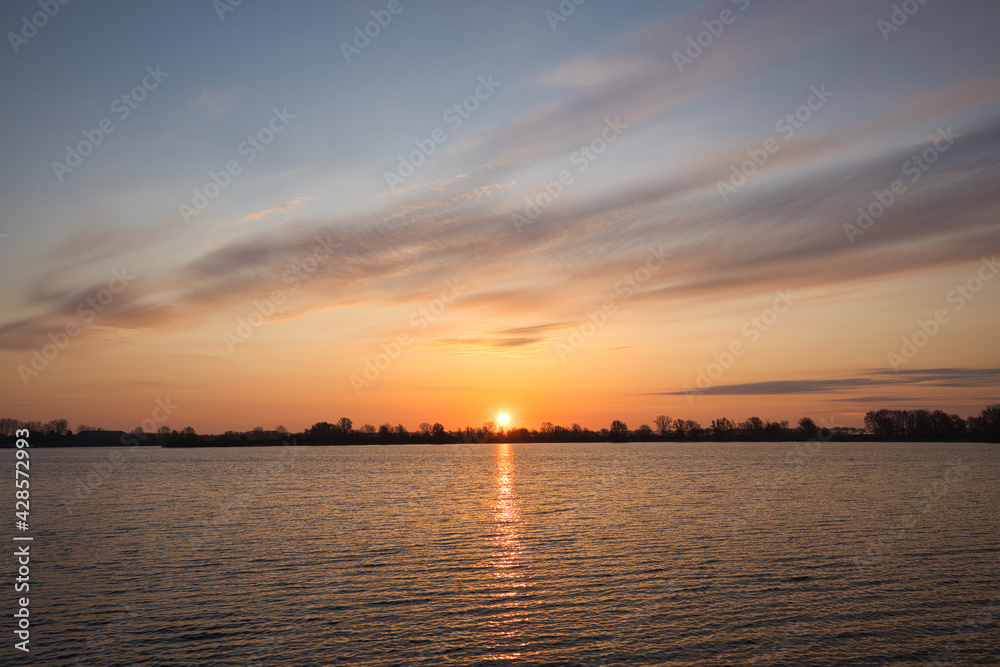 Horizontal view on a lake with a beautiful sunrise, clouds and water. Early morning in spring at “De Zandmeren” in Kerkdriel, Netherlands. With copy space