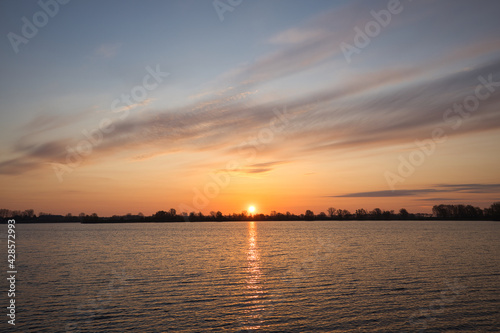 Horizontal view on a lake with a beautiful sunrise, clouds and water. Early morning in spring at “De Zandmeren” in Kerkdriel, Netherlands. With copy space © Tales by Pictures