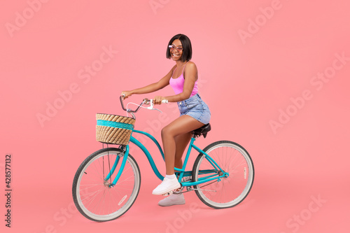 Fototapeta Naklejka Na Ścianę i Meble -  Summer mode. Pretty young black lady in trendy outfit riding bicycle on pink studio background, full length