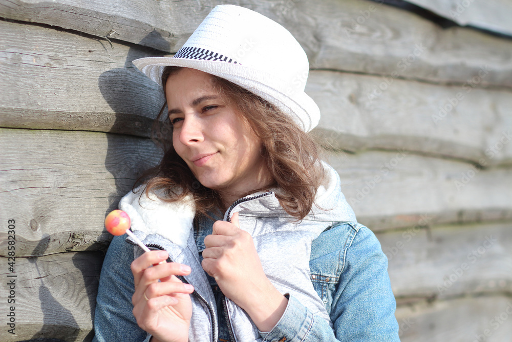 Young pretty woman with Chupa Chups on summer warm day. Beautiful girl in hat and jeans jacket with sweet lollipop.