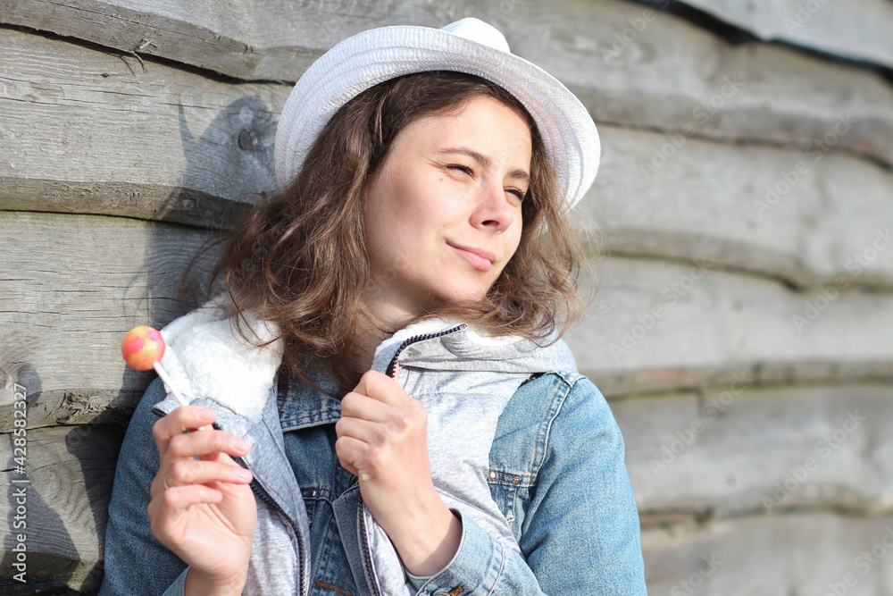 Portrait of dreamy girl with a lollipop near wooden fence. Beautiful young caucasian woman with sweet Chupa-Chups. Spring holidays in ferme.