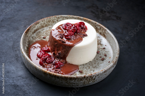 Modern style traditional blancmange almond pudding with cherry and chocolate cream and crumbles served as close-up in a Nordic design plate on a black board