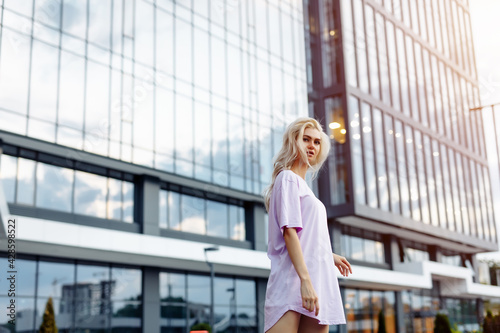 Blissful young woman expressing true emotions while posing on modern street in warm day. Pleased white lady with blonde curls dancing on city buildings background. © Dima