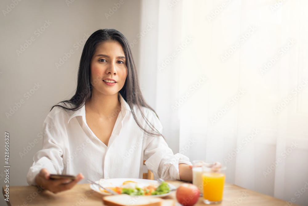 Beautiful woman wearing a white shirt sitting and having a morning breakfast while holding a cell phone. plate of bread, apple, cup of milk. delicious food on dish,  yummy meal. Healthy food concept