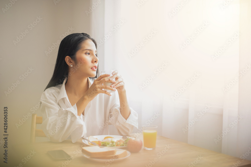 Beautiful woman wearing a white shirt sitting and having a morning breakfast. plate of bread, an apple cup of milk. She looking out the window thinking. Beautiful woman wearing a white shirt sitting and having a morning breakfast. plate of bread, an apple cup of milk. She looking out the window thinking.