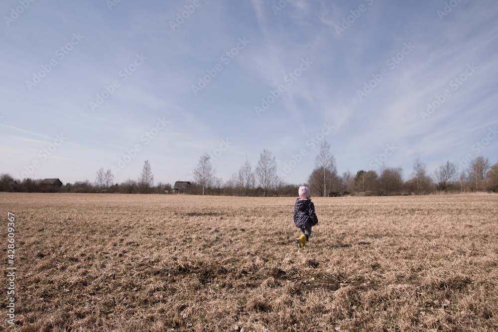 a little girl runs and plays in the field in the spring. a child in rubber boots plays with a stick a little girl runs and plays in the field in the spring. a child in rubber boots plays with a stick