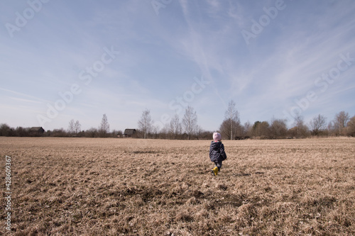 a little girl runs and plays in the field in the spring. a child in rubber boots plays with a stick
