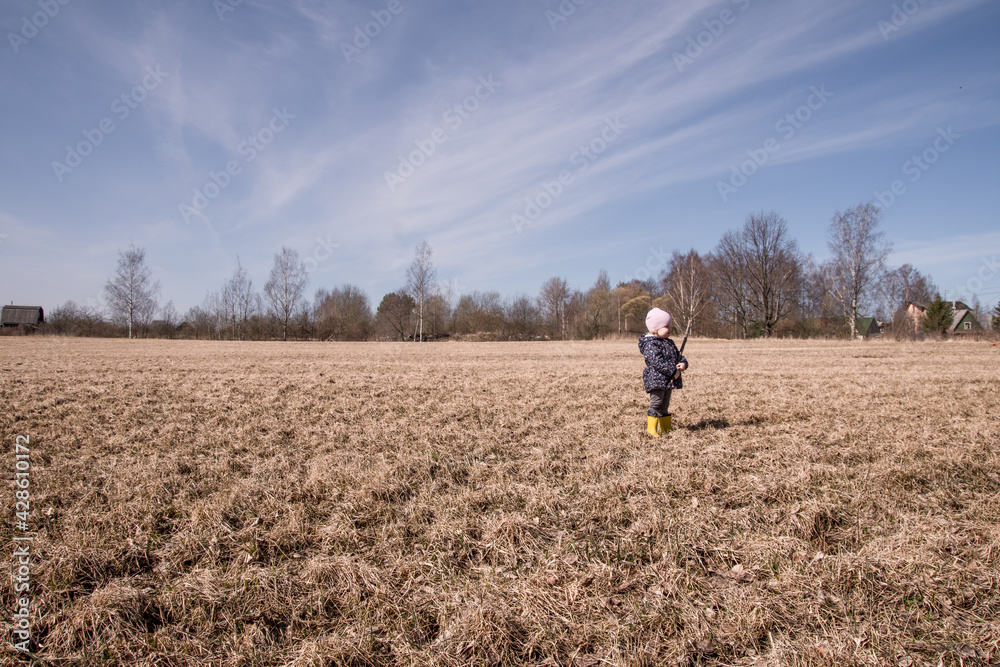 a little girl runs and plays in the field in the spring. a child in rubber boots plays with a stick