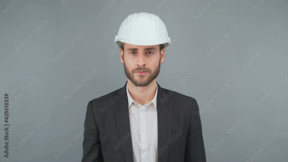 Portrait of a young bearded man in a business suit. Man in white protective hard hat, isolated on gray background. Studio shooting. Portrait of a young bearded man in a business suit. Man in white protective hard hat, isolated on gray background. Studio shooting.