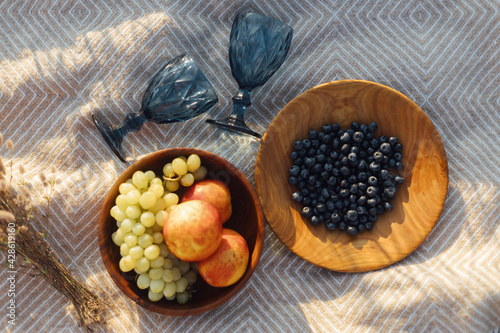Fototapeta Naklejka Na Ścianę i Meble -  Summer picnic. Fresh  fruits in wooden plates and wineglasses on  blanket in sunny light in meadow