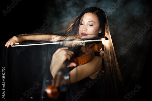 An Asian young woman is playing violin. Violinist is performing on the stage with the veil on her. Emotional playing on the musical instrument. Contour light. Artist.