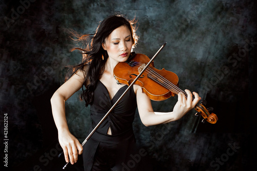 An Asian young woman is playing violin. Violinist is performing on the stage.  Her hair  flying in contour light. Emotional. Musical Instrument
