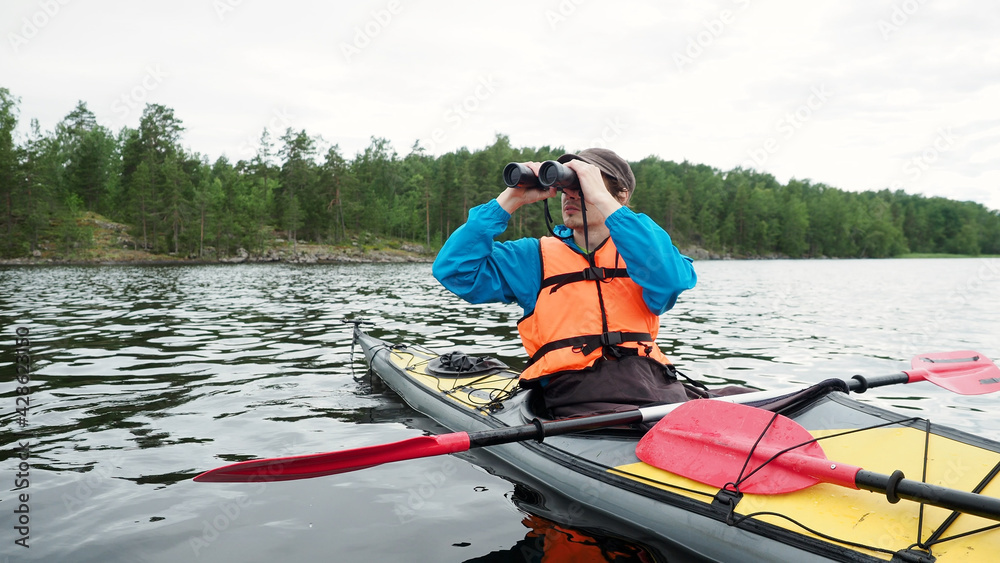 Worried man looks through binoculars sitting in grey yellow kayak boat on summer day. Outdoor activities, water sport concept. Worried man looks through binoculars sitting in grey yellow kayak boat on summer day. Outdoor activities, water sport concept.