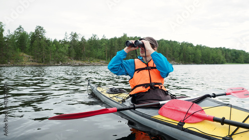 Worried man looks through binoculars sitting in grey yellow kayak boat on summer day. Outdoor activities, water sport concept.