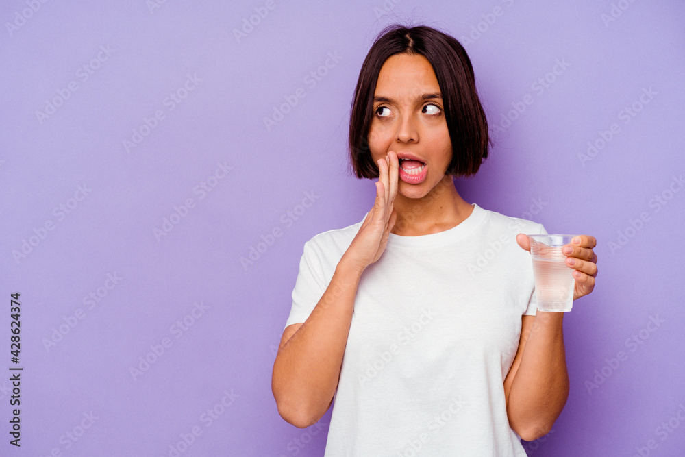 Young mixed race holding a glass of water isolated on purple background is saying a secret hot braking news and looking aside Young mixed race holding a glass of water isolated on purple background is saying a secret hot braking news and looking aside