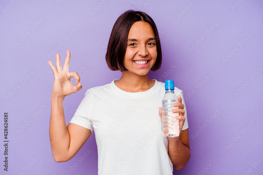 Young mixed race holding a water bottle isolated on purple background cheerful and confident showing ok gesture. Young mixed race holding a water bottle isolated on purple background cheerful and confident showing ok gesture.