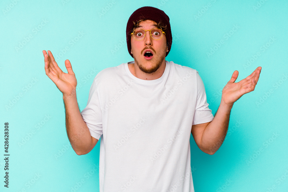 Young caucasian man wearing a wool hat isolated on blue background surprised and shocked. Young caucasian man wearing a wool hat isolated on blue background surprised and shocked.