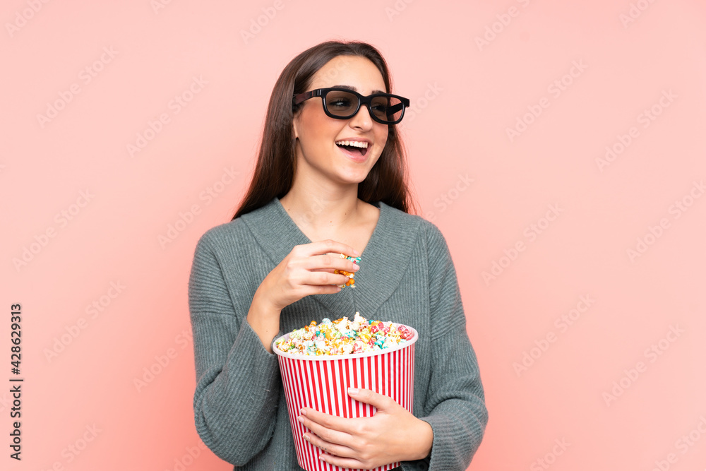 Young caucasian woman isolated on pink background with 3d glasses and holding a big bucket of popcorns