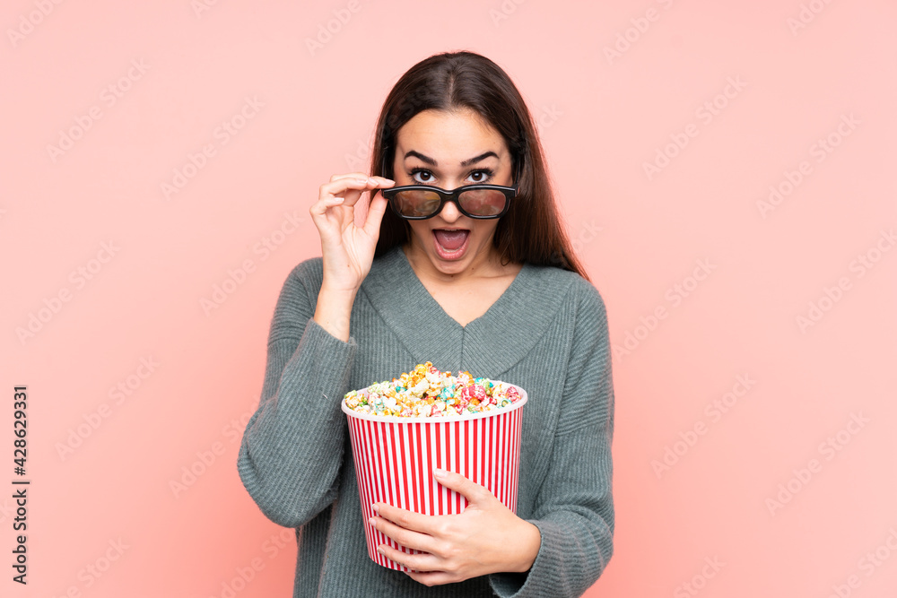 Young caucasian woman isolated on pink background surprised with 3d glasses and holding a big bucket of popcorns