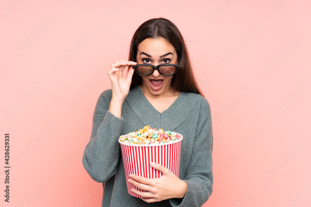 Young caucasian woman isolated on pink background surprised with 3d glasses and holding a big bucket of popcorns Young caucasian woman isolated on pink background surprised with 3d glasses and holding a big bucket of popcorns