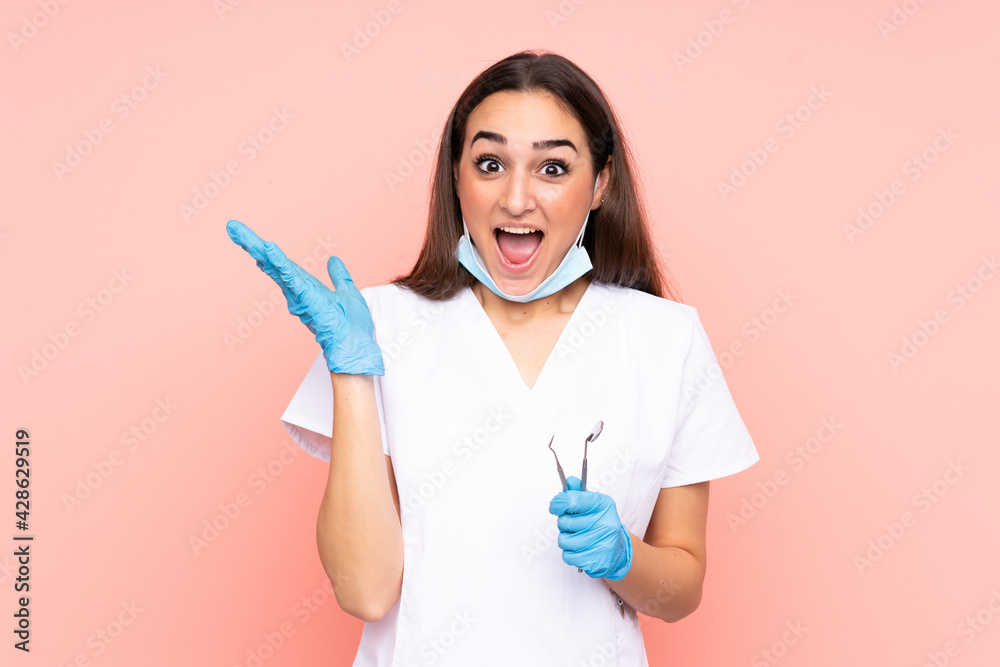 Woman dentist holding tools isolated on pink background with surprise facial expression Woman dentist holding tools isolated on pink background with surprise facial expression
