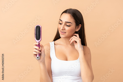 Young caucasian woman isolated on beige background with hair comb and looking it