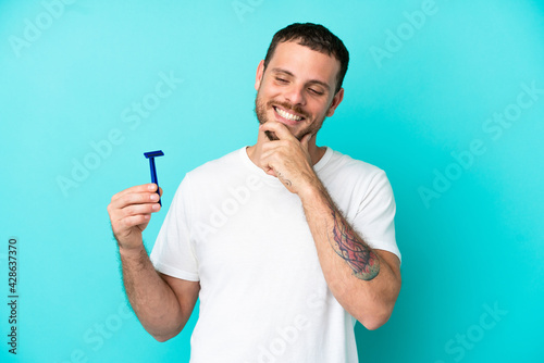 Brazilian man shaving his beard isolated on blue background looking to the side and smiling