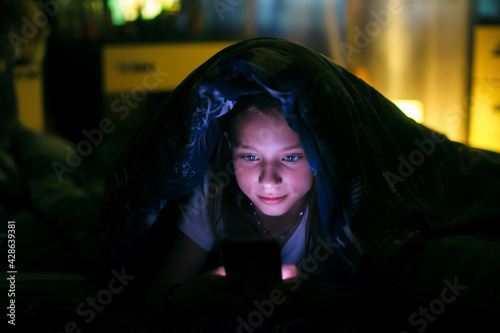 Little girl at dark looks at smartphone under a blanket in bed.