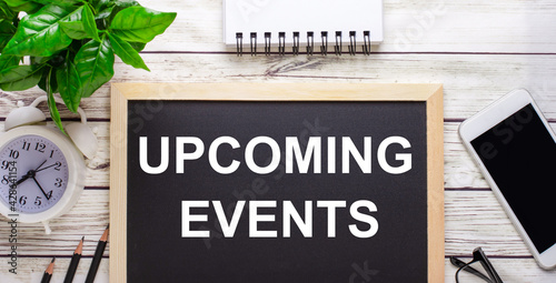 UPCOMING EVENTS written on a black background near pencils, a smartphone, a white notepad and a green plant in a pot