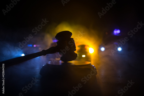 Law theme, mallet of the judge on wooden desk . Silhouette of police cars on backside. Law gavel on dark foggy background with light