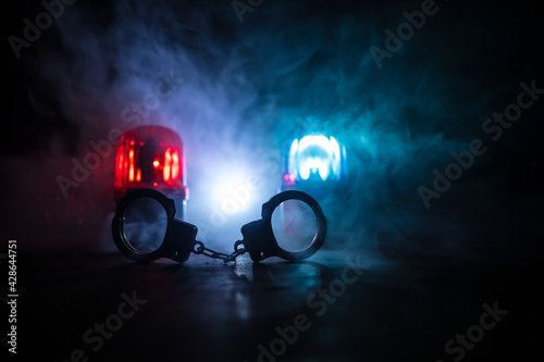 Police raid at night and you are under arrest concept. Silhouette of handcuffs with police car on backside. Image with the flashing red and blue police lights at foggy background. © zef art