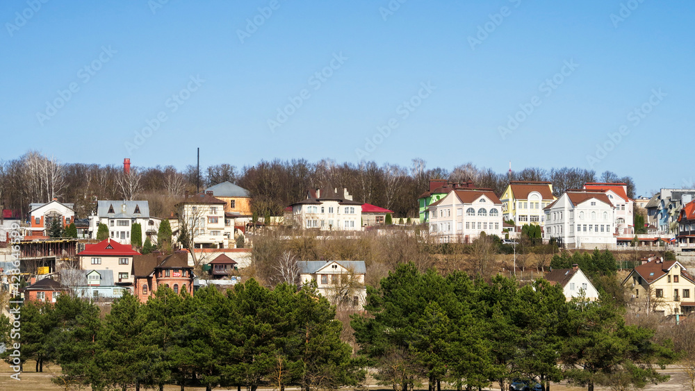 Village houses on a hill against a blue sky. Meadow with trees and grass in the field in front of the house. Horizontal village landscape with summer sunny day.