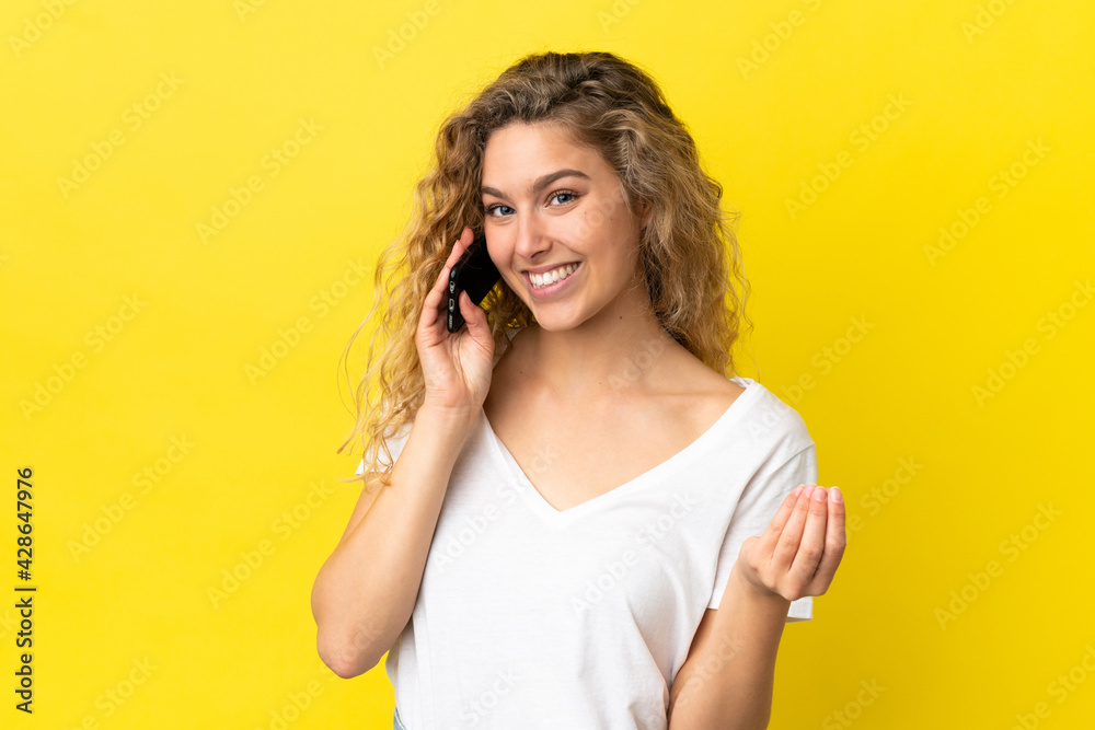 Young blonde woman using mobile phone isolated on yellow background making money gesture Young blonde woman using mobile phone isolated on yellow background making money gesture