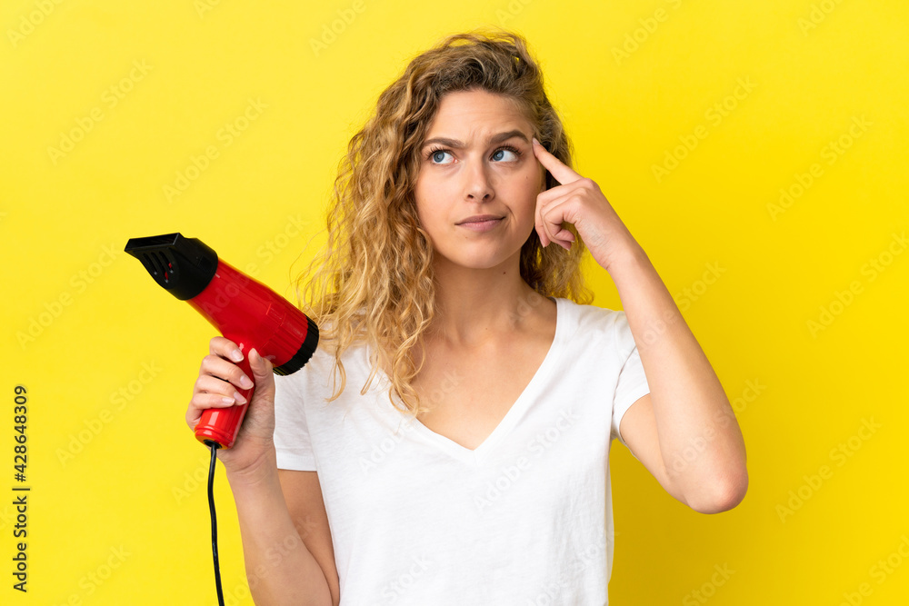 Young blonde woman holding a hairdryer isolated on yellow background having doubts and thinking Young blonde woman holding a hairdryer isolated on yellow background having doubts and thinking