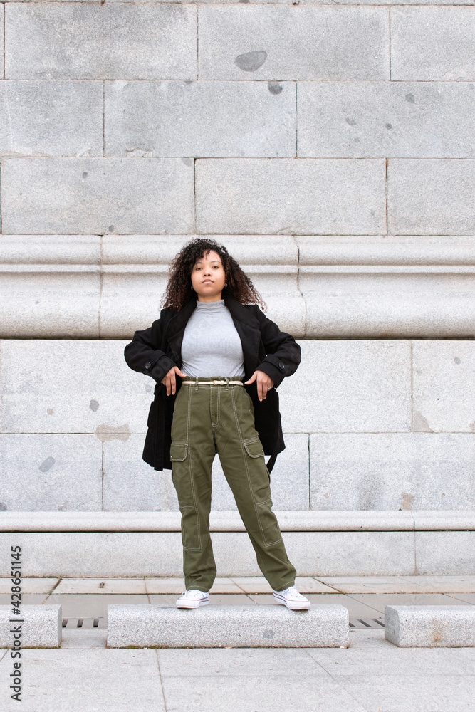 Young woman with curly hair looking directly into the camera. Black jacket and green pants. Vertical photo.