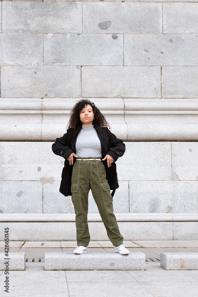 Young woman with curly hair looking directly into the camera. Black jacket and green pants. Vertical photo. Young woman with curly hair looking directly into the camera. Black jacket and green pants. Vertical photo.
