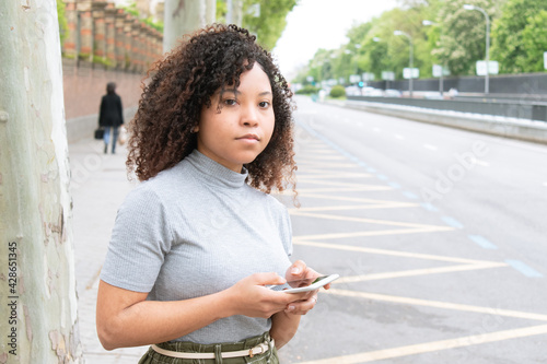 young black girl with curly hair in the city waiting for a cab or uber with cell phone in hand.