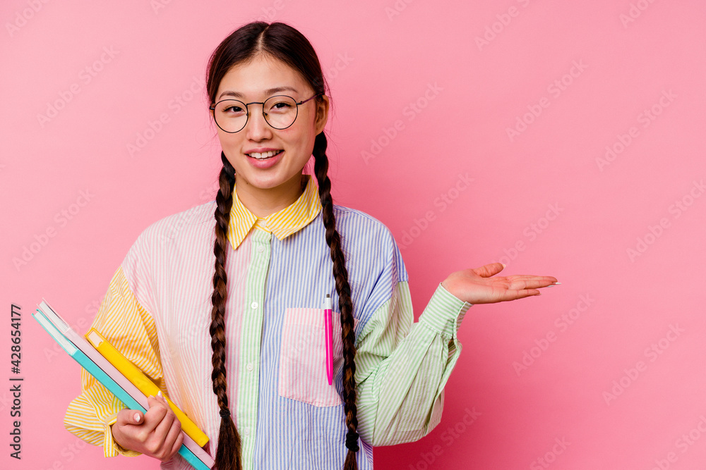 Young chinese student woman holding books wearing a fashion multicolour shirt and braid, isolated on pink background showing a copy space on a palm and holding another hand on waist. Young chinese student woman holding books wearing a fashion multicolour shirt and braid, isolated on pink background showing a copy space on a palm and holding another hand on waist.
